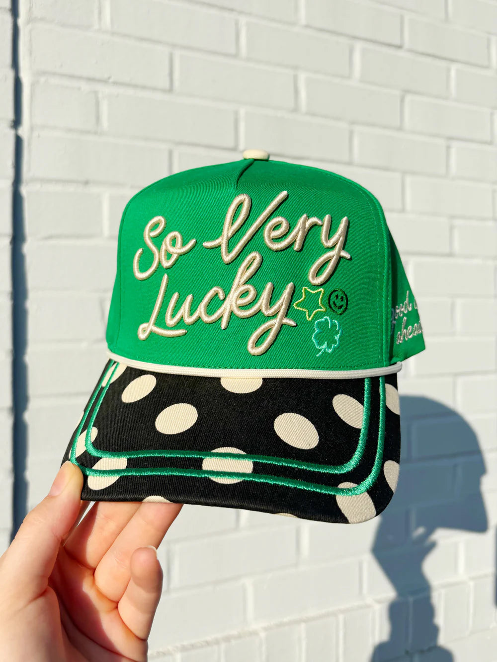 Green cap with 'So Very Lucky' text held against a white brick wall