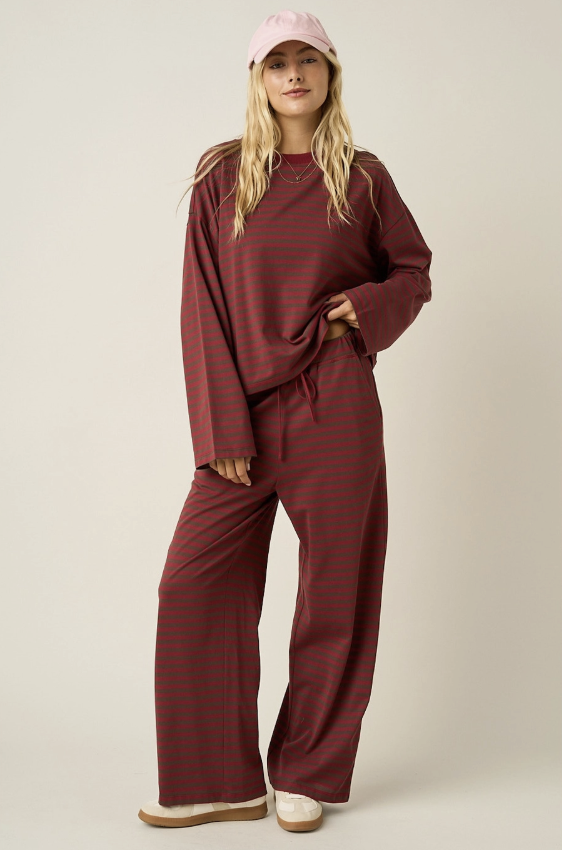 A woman modeling a matching two-piece loungewear set consisting of a long-sleeve crewneck top and wide-leg pants, both in a dark red and dark green striped pattern. She is also wearing a light pink baseball cap and cream-colored shoes, standing against a solid light beige studio background.