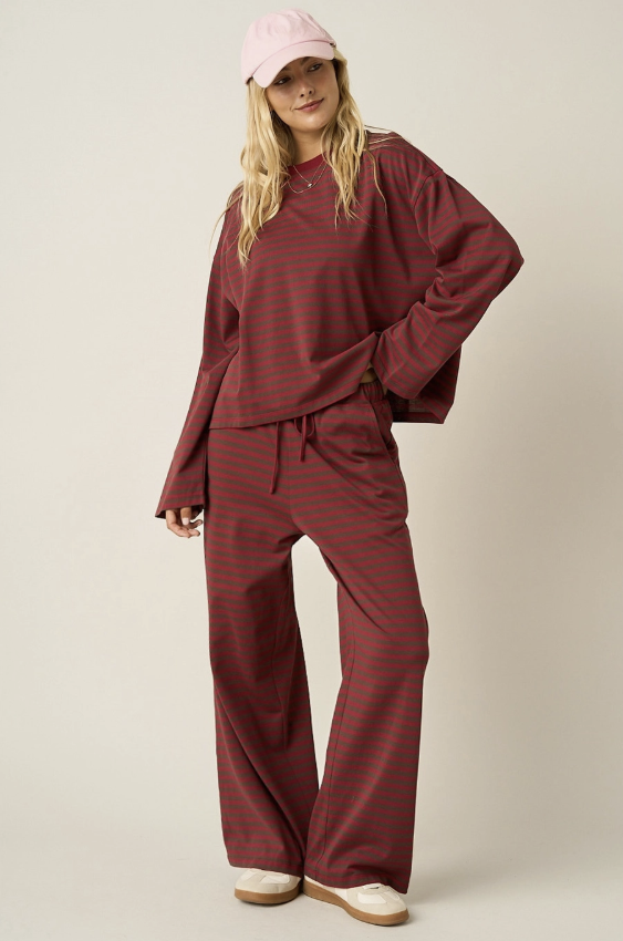 A woman modeling a matching two-piece loungewear set consisting of a long-sleeve crewneck top and wide-leg pants, both in a dark red and dark green striped pattern. She is also wearing a light pink baseball cap and beige sneakers against a plain beige studio background.