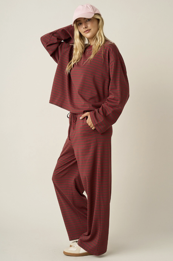 A woman modeling a matching two-piece lounge set consisting of a cropped long-sleeve top and wide-leg pants, both in a dark red and green striped pattern. She is also wearing a light pink baseball cap and off-white sneakers against a plain beige studio background.