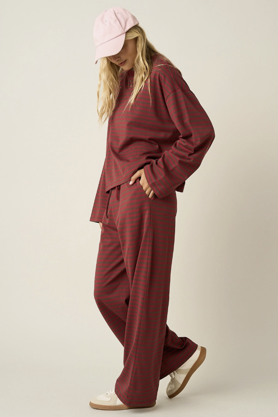 A woman modeling a matching burgundy and dark red striped lounge set, consisting of a long-sleeve top and wide-leg pants, styled with a light pink baseball cap and white slip-on shoes against a neutral studio background.
