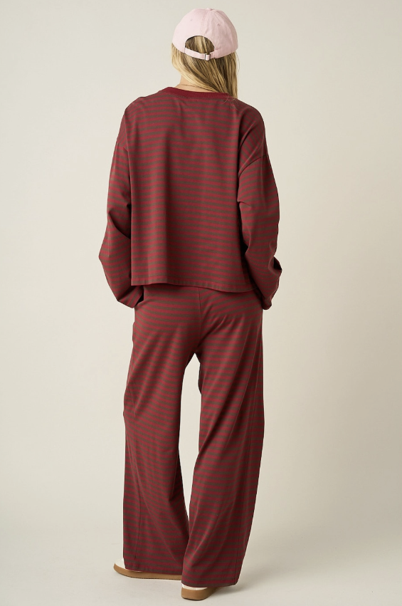 A model shown from the back wearing a matching two-piece loungewear set consisting of a long-sleeve cropped top and wide-leg pants, both in a dark red and dark green striped pattern. The model is also wearing a light pink baseball cap and tan slippers, set against a plain light beige studio background.