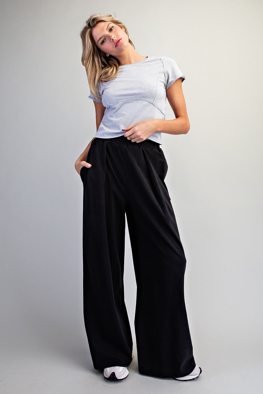 A woman modeling a light blue short-sleeve t-shirt and black wide-leg pants against a neutral gray studio background.