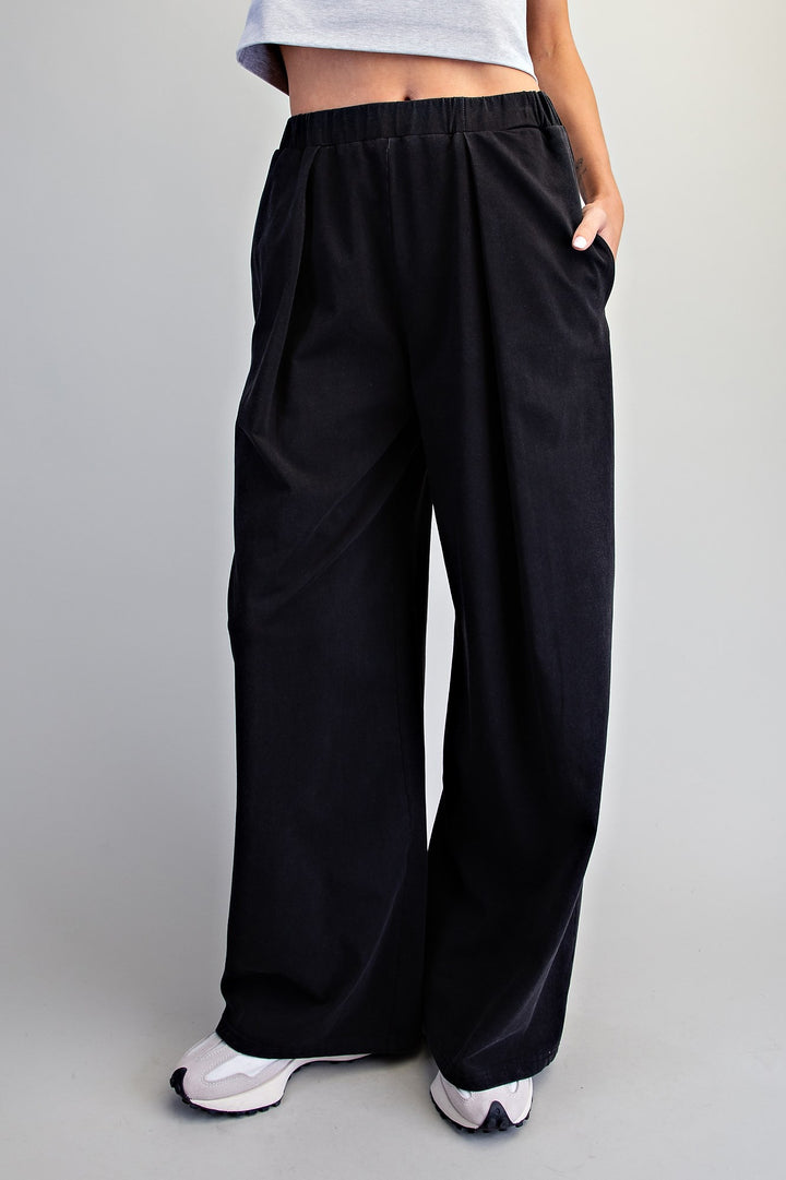 A person wearing loose black wide-leg pants with an elastic waistband and side pockets, paired with white sneakers, shown from the waist down against a light gray studio background.