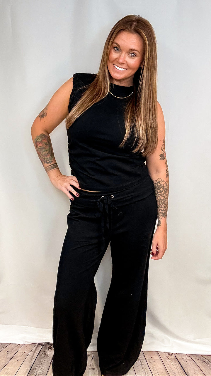 A woman modeling a black sleeveless mock neck top and matching black wide-leg drawstring pants against a white studio backdrop.