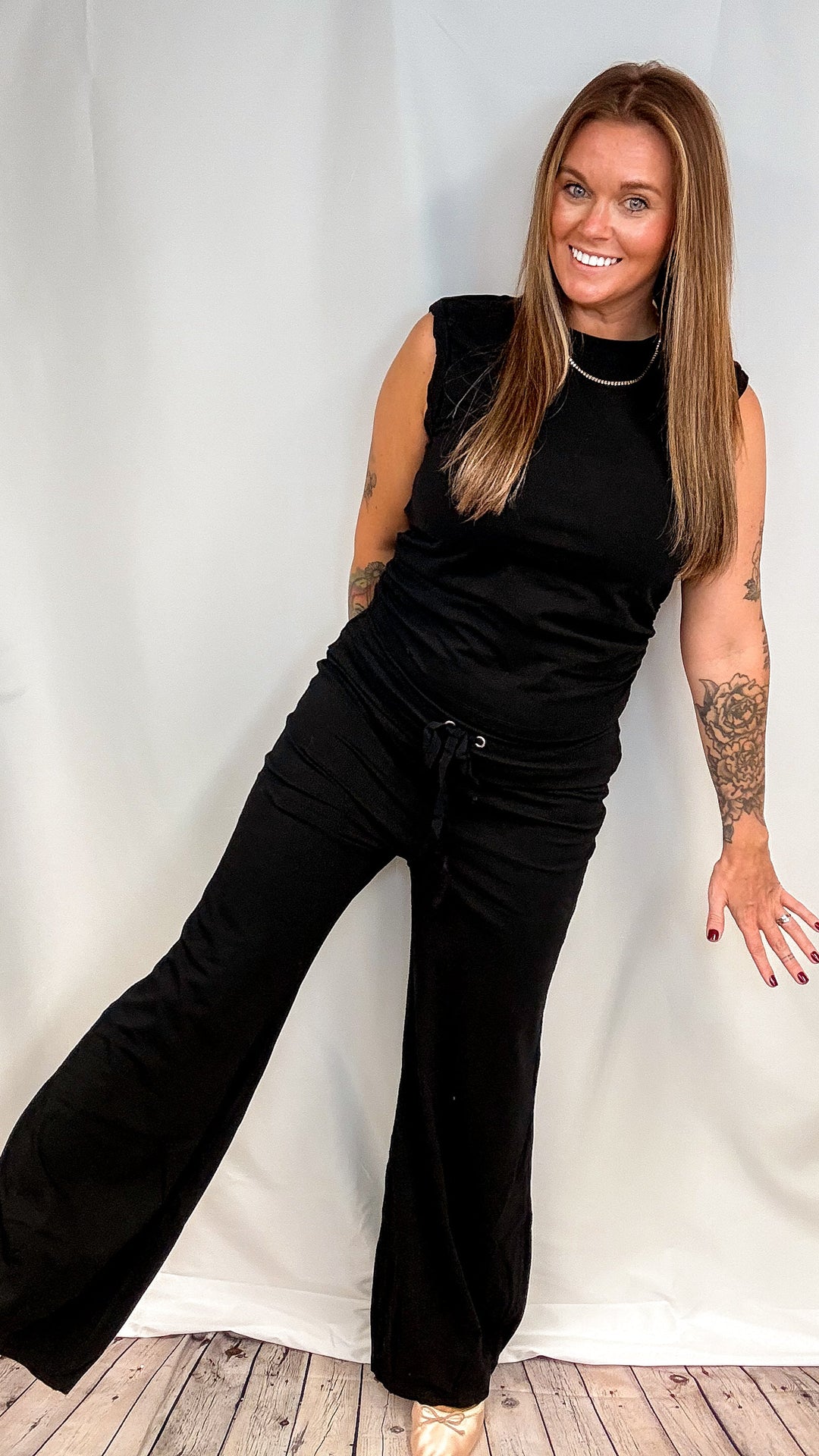 A woman modeling a black sleeveless top and matching black wide-leg pants with a drawstring waist, standing against a white fabric backdrop on a wood floor.