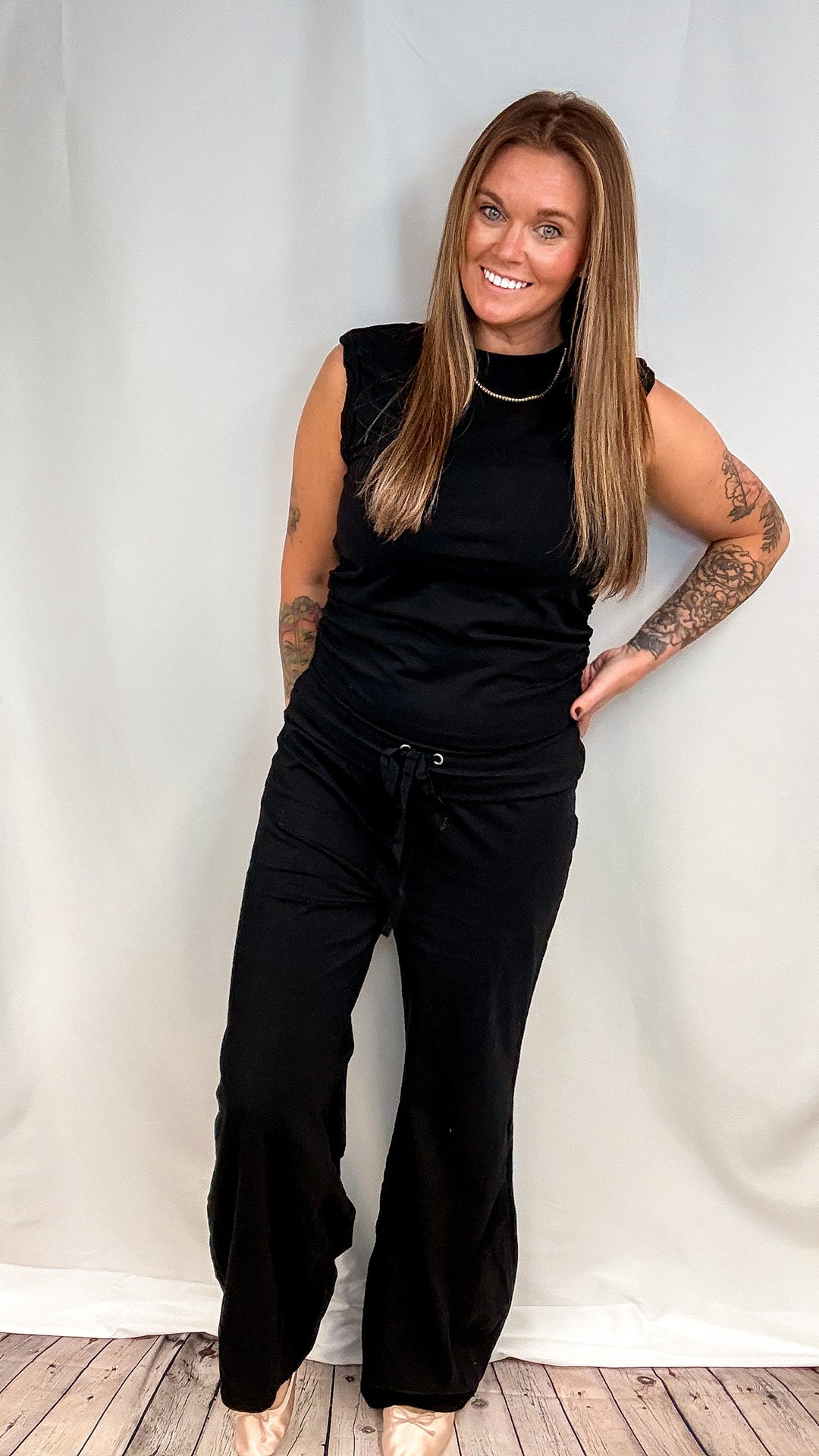 A woman modeling a black sleeveless top and black wide-leg drawstring pants against a white fabric backdrop.