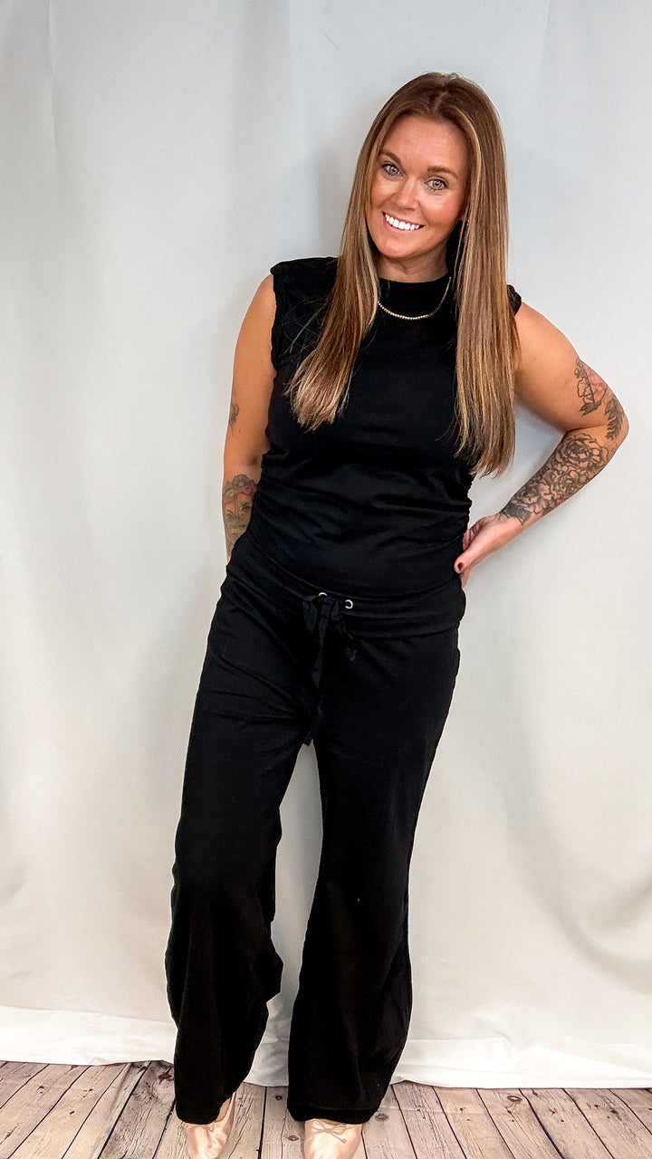 A woman modeling a black sleeveless top and black wide-leg drawstring pants against a white fabric backdrop.