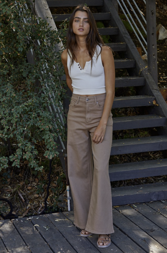 A woman stands on outdoor wooden stairs wearing a white sleeveless crop top and high-waisted, wide-leg taupe pants, paired with flat sandals.