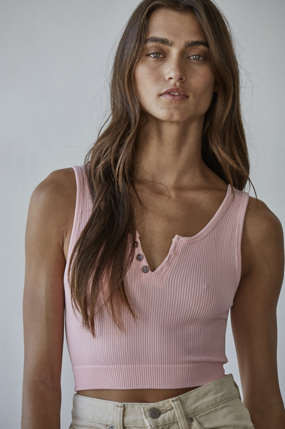 A woman wearing a light pink ribbed knit crop top with a split henley neckline and decorative buttons, paired with light beige pants, against a neutral gray studio background.