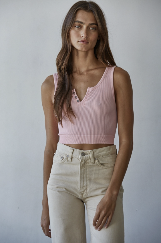 A woman modeling a light pink ribbed knit Henley crop top with three decorative buttons, paired with cream-colored jeans, against a plain light gray wall.
