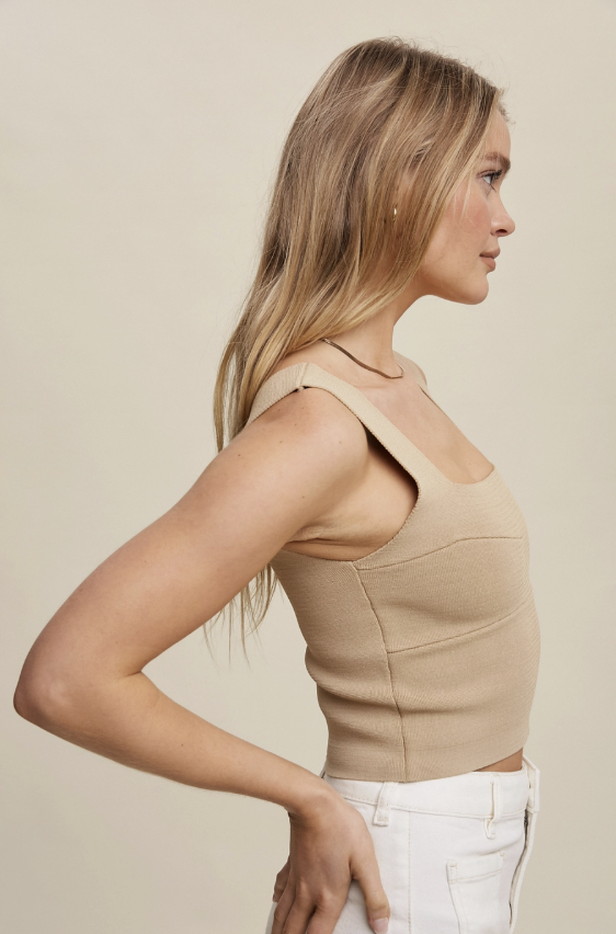 A woman in side profile wearing a beige ribbed knit crop top with wide straps and white pants, set against a plain beige studio background.