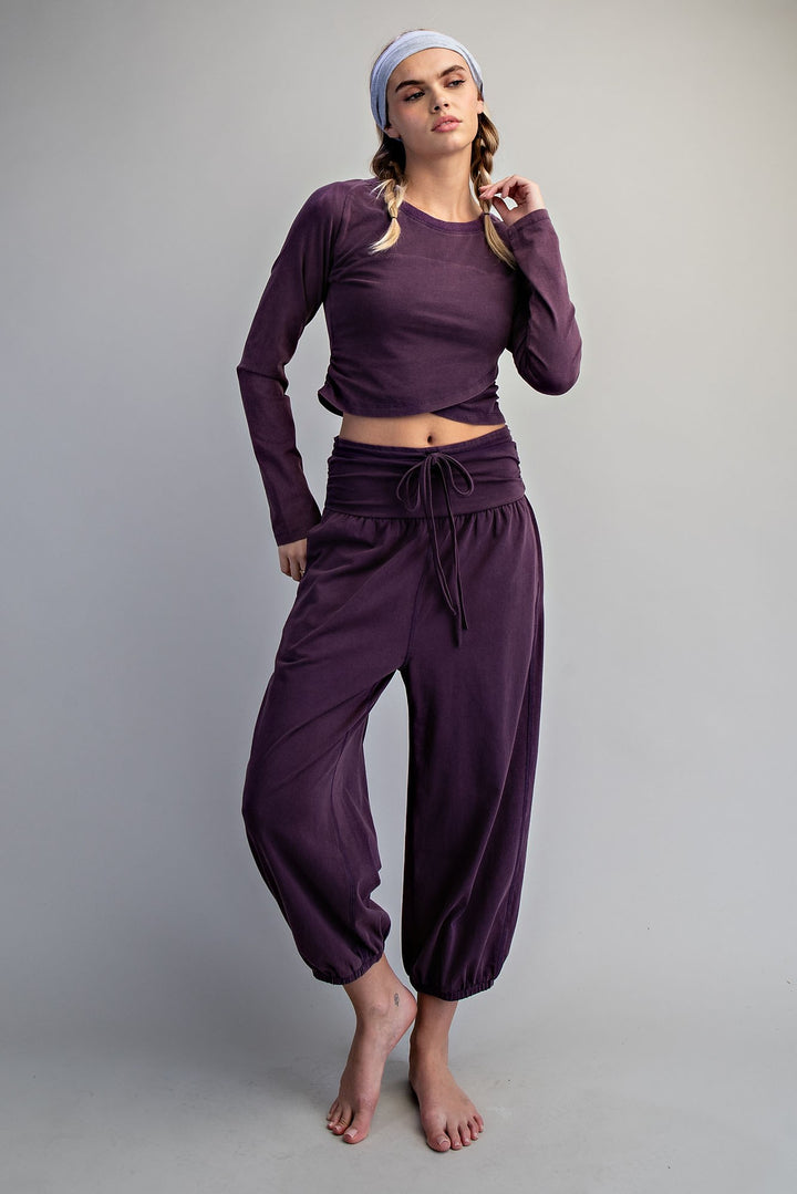 A woman modeling a matching dark purple loungewear set consisting of a long-sleeve crop top and high-waisted drawstring pants, paired with a light blue headband, against a plain gray studio background.