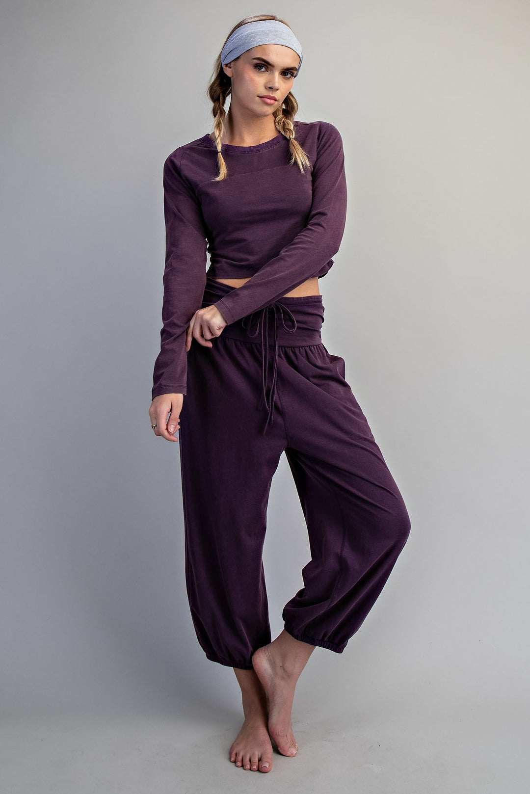 A woman modeling a matching deep purple loungewear set consisting of a long-sleeve crop top and high-waisted drawstring sweatpants, paired with a light blue headband, against a neutral gray studio background.