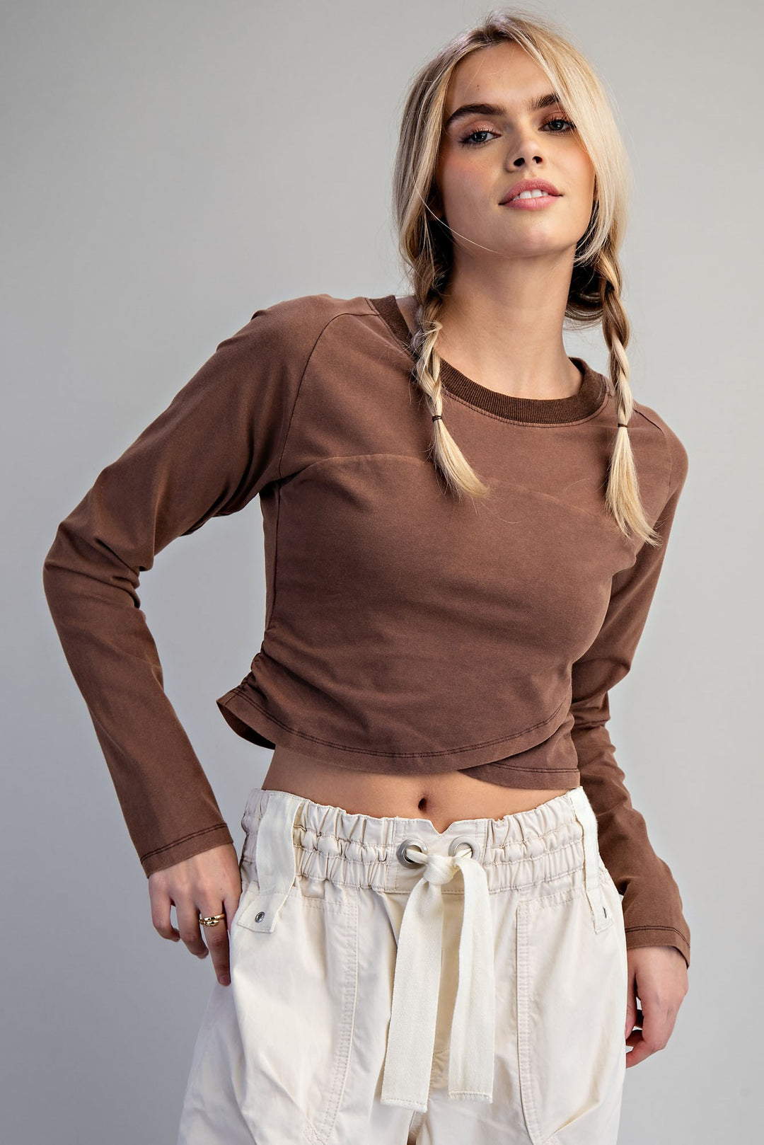 A blonde model with braided hair wearing a brown long-sleeve crop top and cream drawstring pants, photographed against a solid gray studio background.