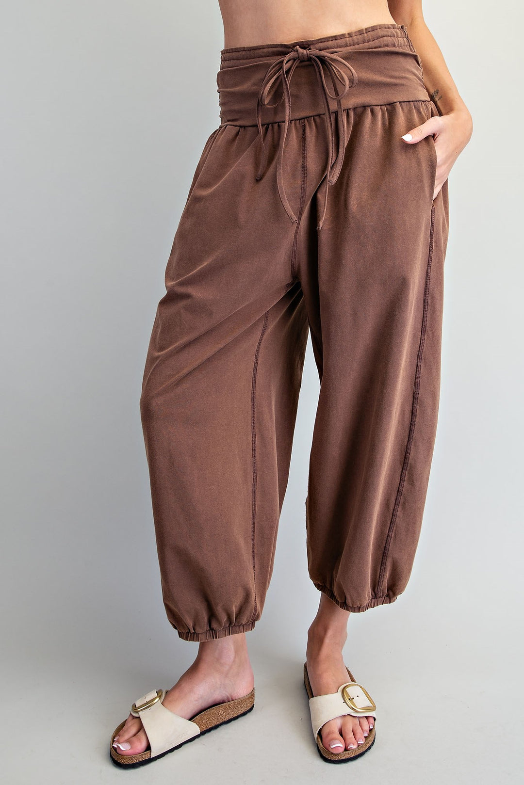 A person wearing loose brown harem-style pants with a drawstring waist and elastic cuffs, paired with beige slide sandals, against a plain light gray studio background.
