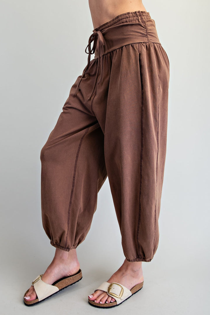 A person wearing loose brown harem-style pants with a drawstring waist and elastic cuffs, paired with beige slide sandals, photographed against a plain light gray studio background.