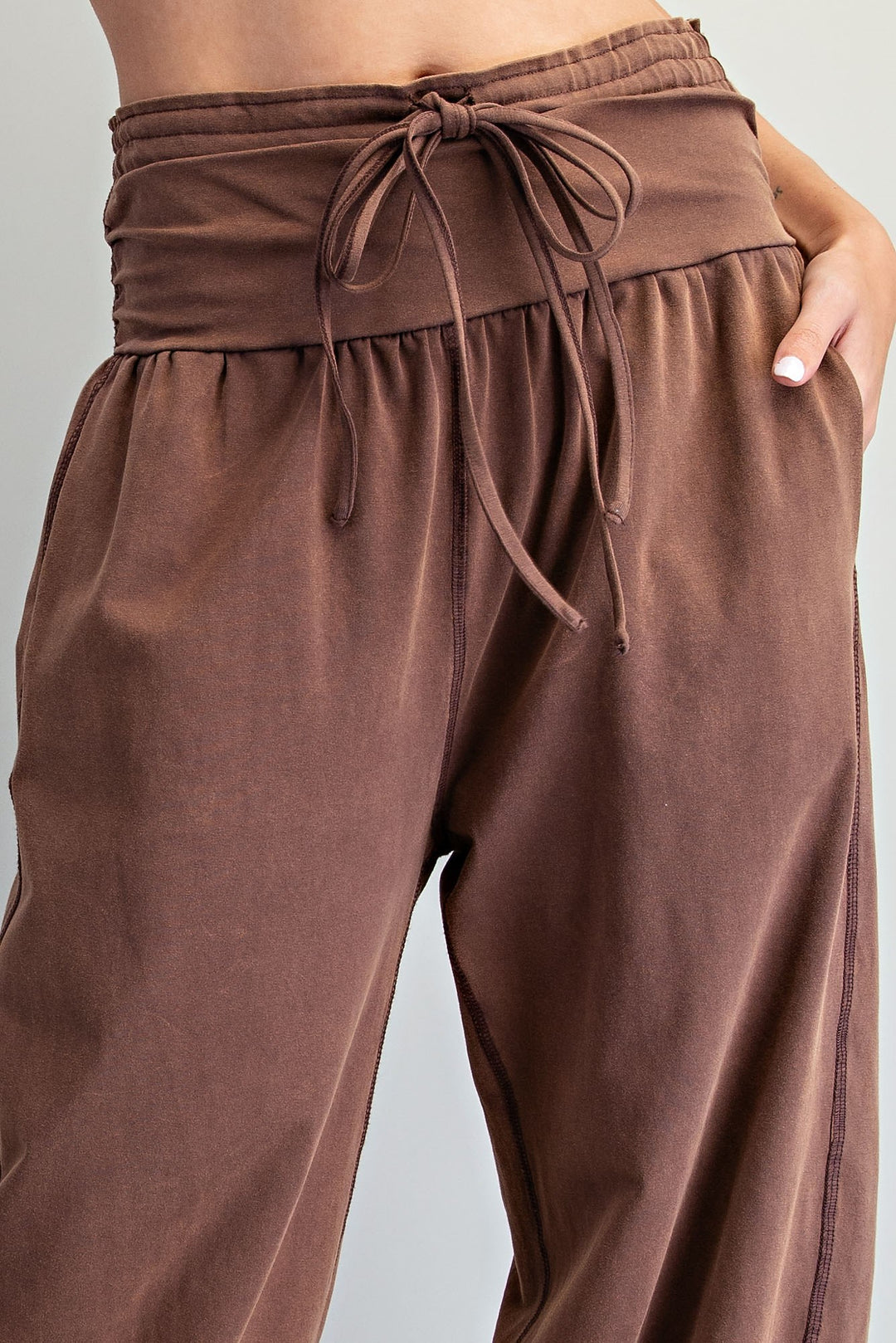 Close-up of a person wearing loose brown drawstring pants with a wide elastic waistband and side pockets, shown against a light gray studio background.