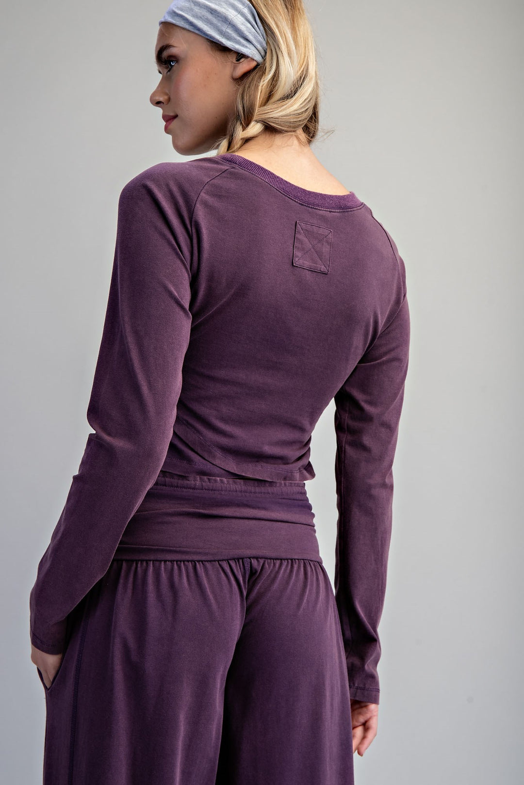 A woman shown from the back wearing a matching purple long-sleeve top and wide-leg pants set, with a light blue and white striped headband, against a plain gray studio background.