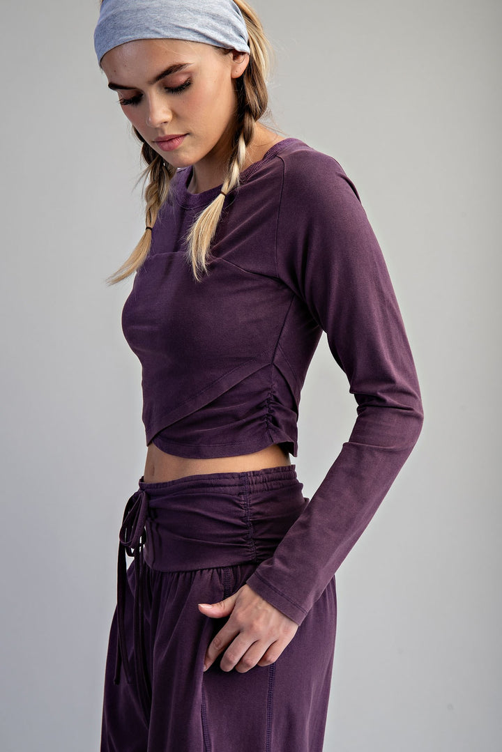 A woman wearing a matching purple two-piece lounge set, featuring a long-sleeve crop top with ruching and matching high-waisted drawstring pants, paired with a light blue headband against a neutral gray background.
