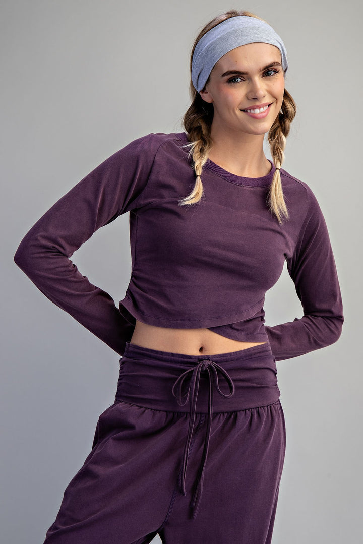 A woman modeling a matching athletic loungewear set in deep purple, featuring a long-sleeve cropped top and high-waisted drawstring pants, paired with a light blue headband against a solid gray background.