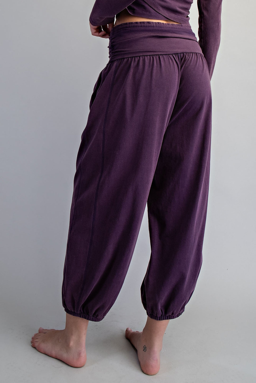 A person wearing loose, high-waisted harem-style pants in a deep plum purple, shown from the back against a plain light gray studio background.