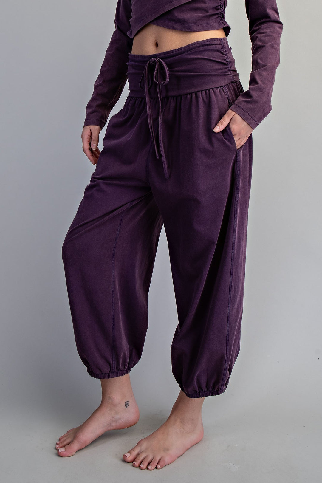 A person wearing loose, high-waisted harem-style pants in a deep plum purple, paired with a matching long-sleeve cropped top. The pants feature a drawstring waist, side pockets, and gathered elastic cuffs at the ankles, shown against a plain light gray studio background.