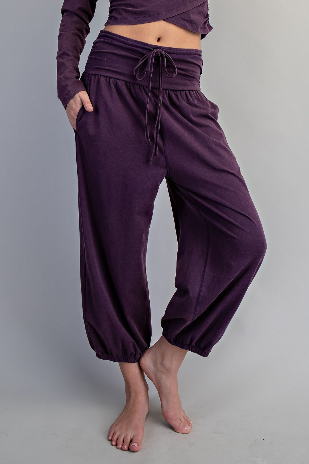 A person wearing loose, high-waisted harem-style pants in a deep purple or eggplant shade, paired with a matching long-sleeve crop top, shown against a plain light gray studio background.