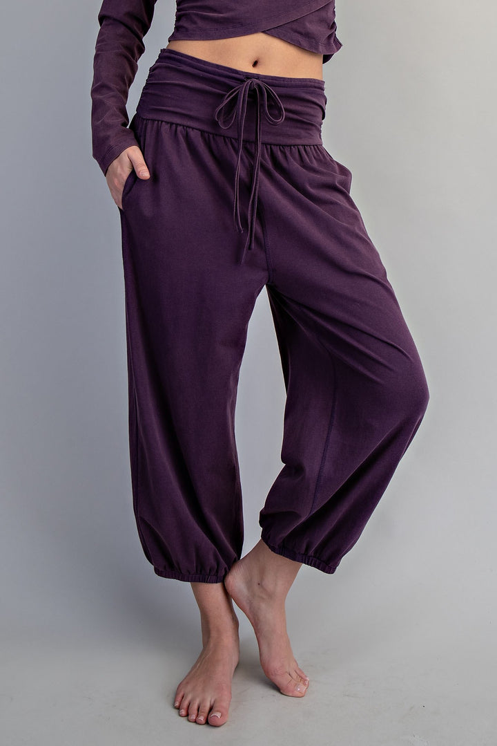 A person wearing loose, high-waisted harem-style pants in a deep purple or eggplant shade, paired with a matching long-sleeve crop top, shown against a plain light gray studio background.