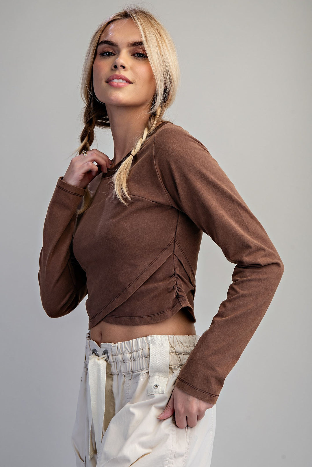 A blonde model with braided hair wearing a brown long-sleeve crop top with side ruching and cream-colored pants, posing against a solid light gray background.