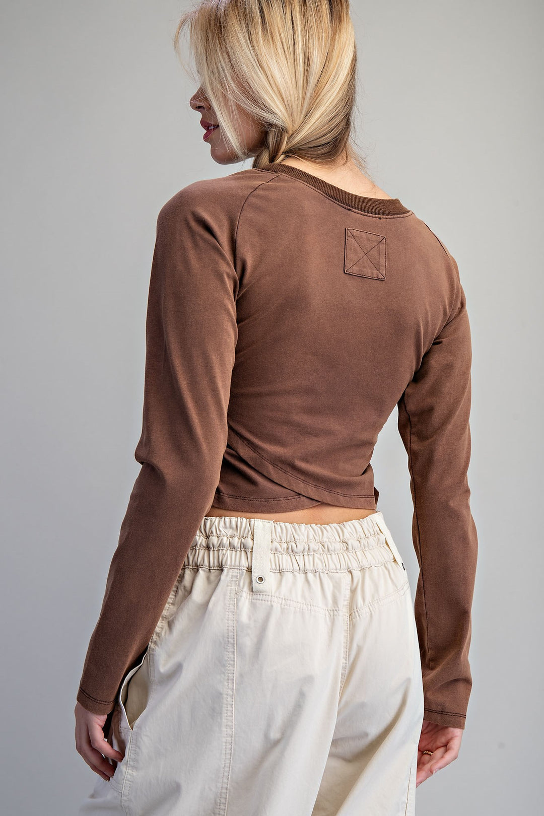 A woman shown from the back wearing a brown long-sleeve crop top and cream-colored pants against a solid gray studio background.