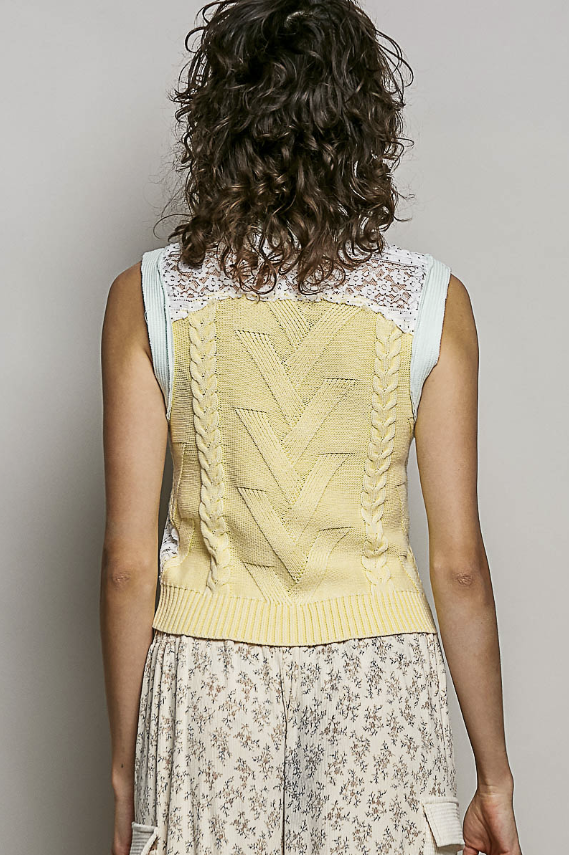 Person wearing a light yellow knitted vest over a white lace top with floral pants on a plain background