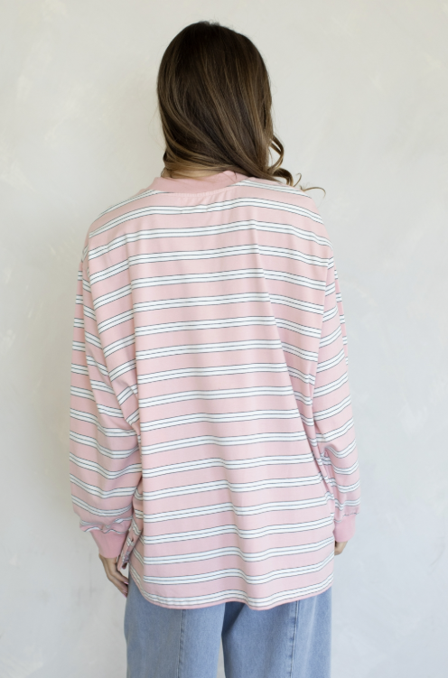 A woman viewed from behind wearing a pink long-sleeve striped shirt with thin white and gray horizontal lines, paired with light blue jeans, against a light textured wall.