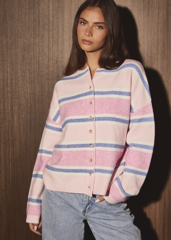 A woman wearing a pink, white, and blue striped knit cardigan with gold buttons, paired with light-wash blue jeans, standing against a dark wood-paneled wall.