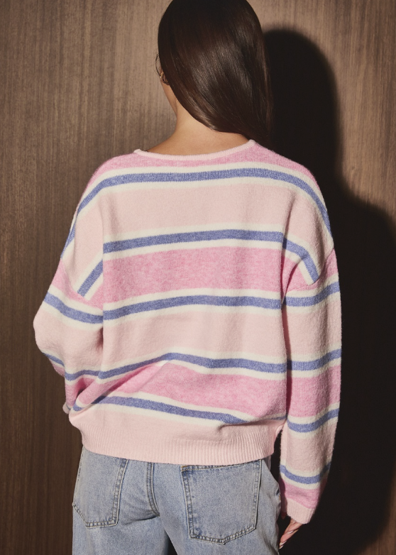 A woman viewed from behind wearing a pink knit sweater with horizontal blue and white stripes, paired with light-wash blue jeans, standing against a dark wood-paneled wall.