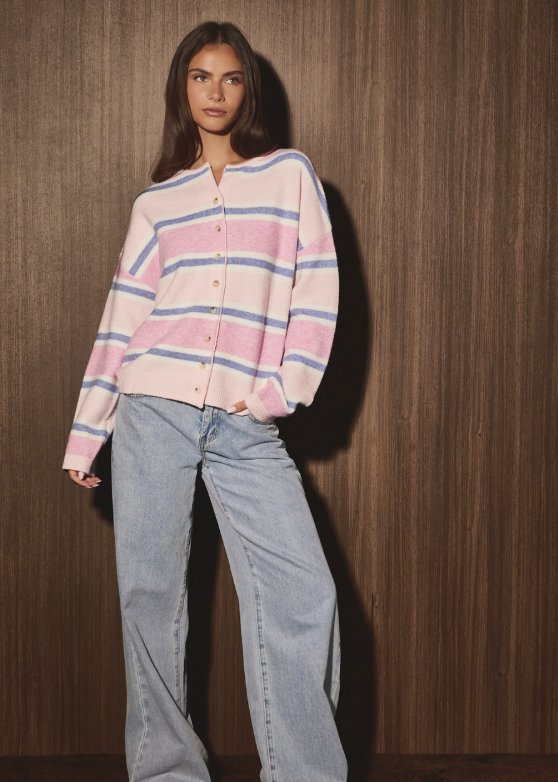 A woman stands against a dark wood-paneled wall wearing a pink, light blue, and white striped button-up cardigan with wide-leg light-wash jeans and off-white sneakers.