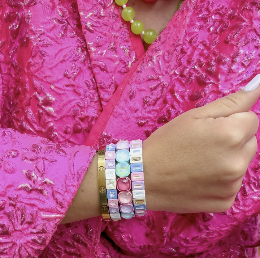A close-up of a person's wrist wearing a stack of pastel beaded bracelets, including a gold-toned bracelet with small round charms, a light blue bracelet with oval beads, a pink bracelet with letter beads, and a white bracelet with pastel square beads. The person is wearing a bright pink textured top.
