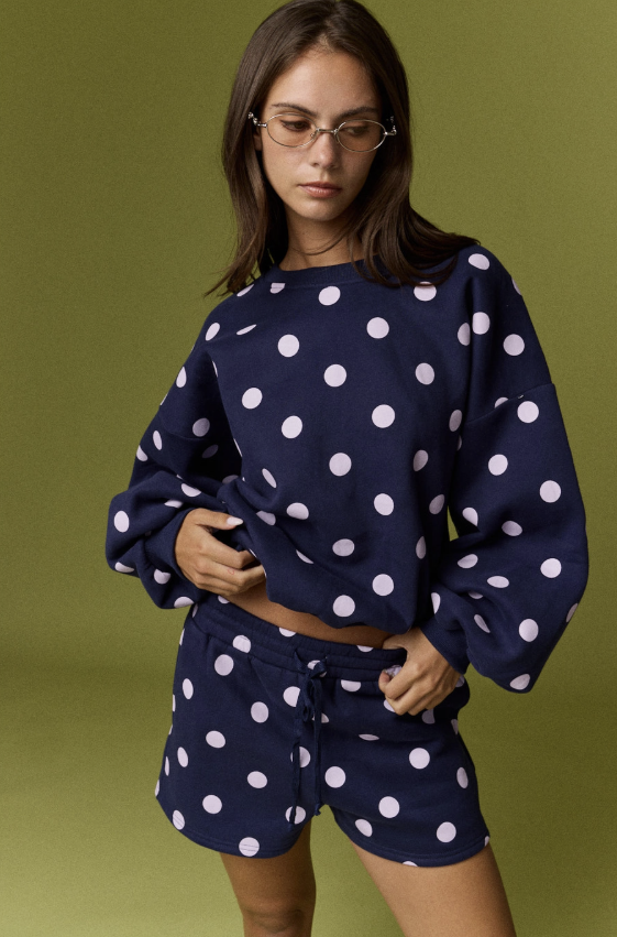 A woman wearing a navy blue and white polka dot lounge set, consisting of a cropped long-sleeve sweatshirt and matching drawstring shorts, posed against a solid olive green background.