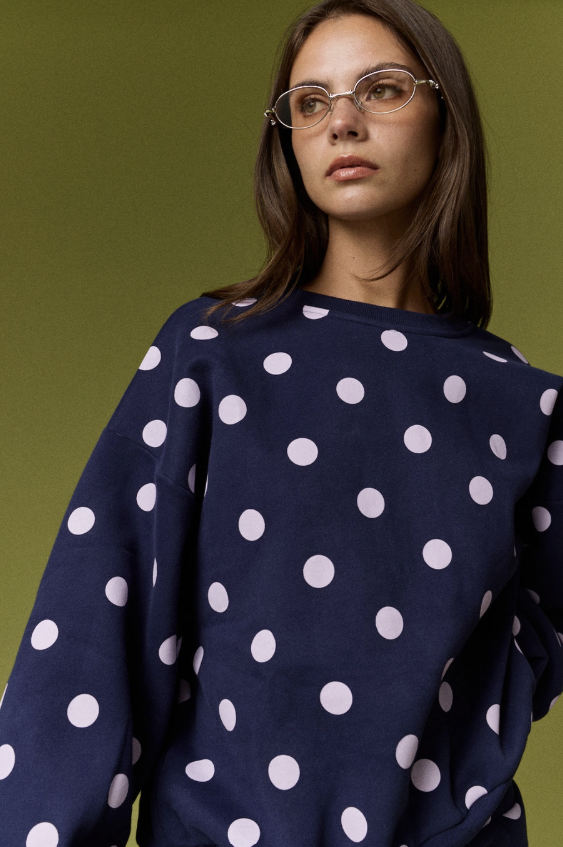 A woman wearing a navy blue sweatshirt with a white polka dot pattern, photographed against a solid olive green background.