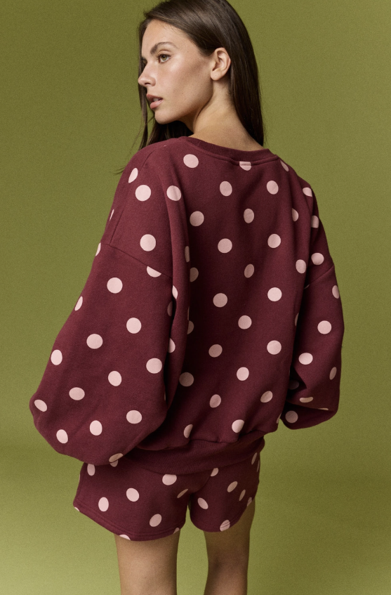 A woman shown from the back wearing a burgundy long-sleeve sweatshirt and matching shorts set, both covered in a light pink polka dot pattern, against an olive green studio background.