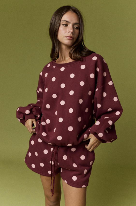 A woman modeling a matching burgundy and pink polka-dot lounge set, featuring a long-sleeve top with gathered cuffs and matching shorts, set against a solid olive green background.