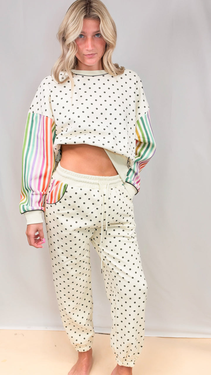Woman wearing matching cream sweatsuit with rainbow color stripe sleeves and black polka dots on plain background