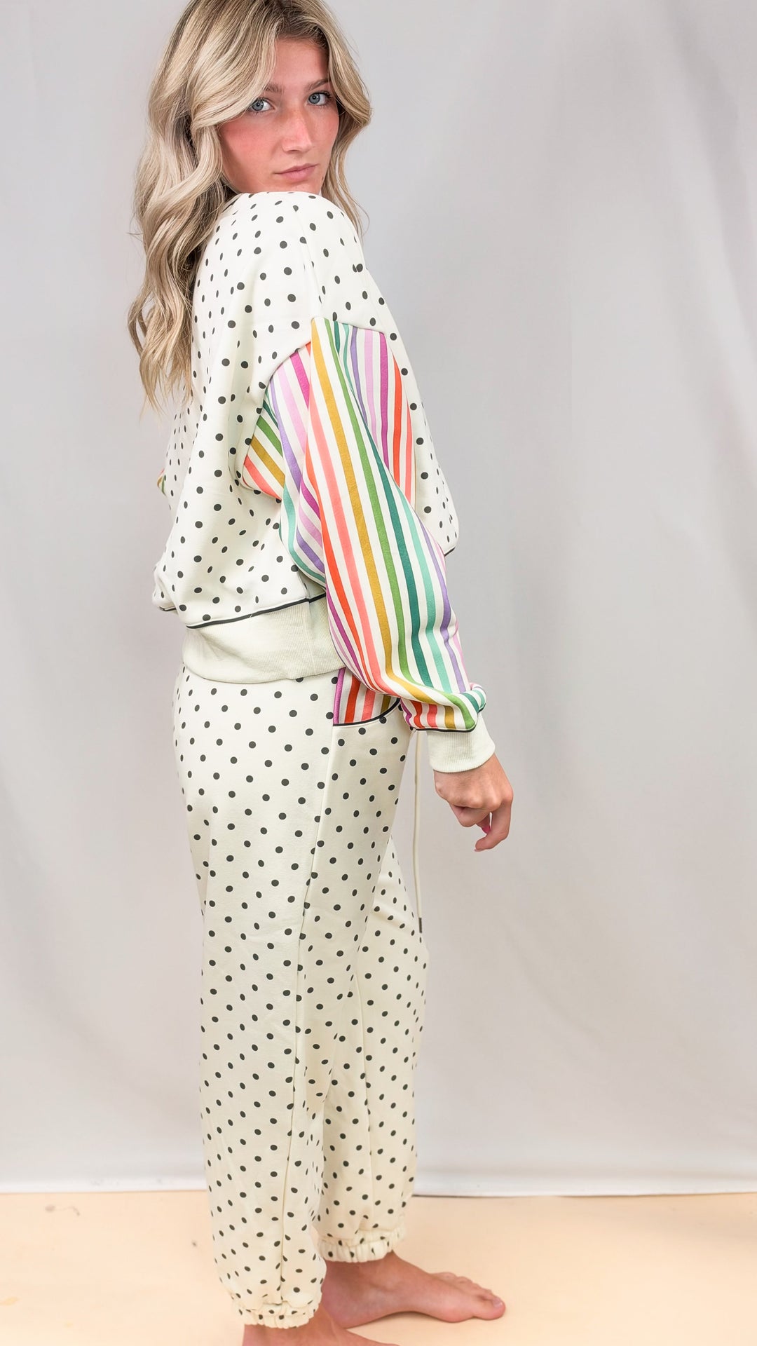 Woman shown from side view wearing cream matching sweatsuit with rainbow color stripe sleeves and black polka dots on plain background