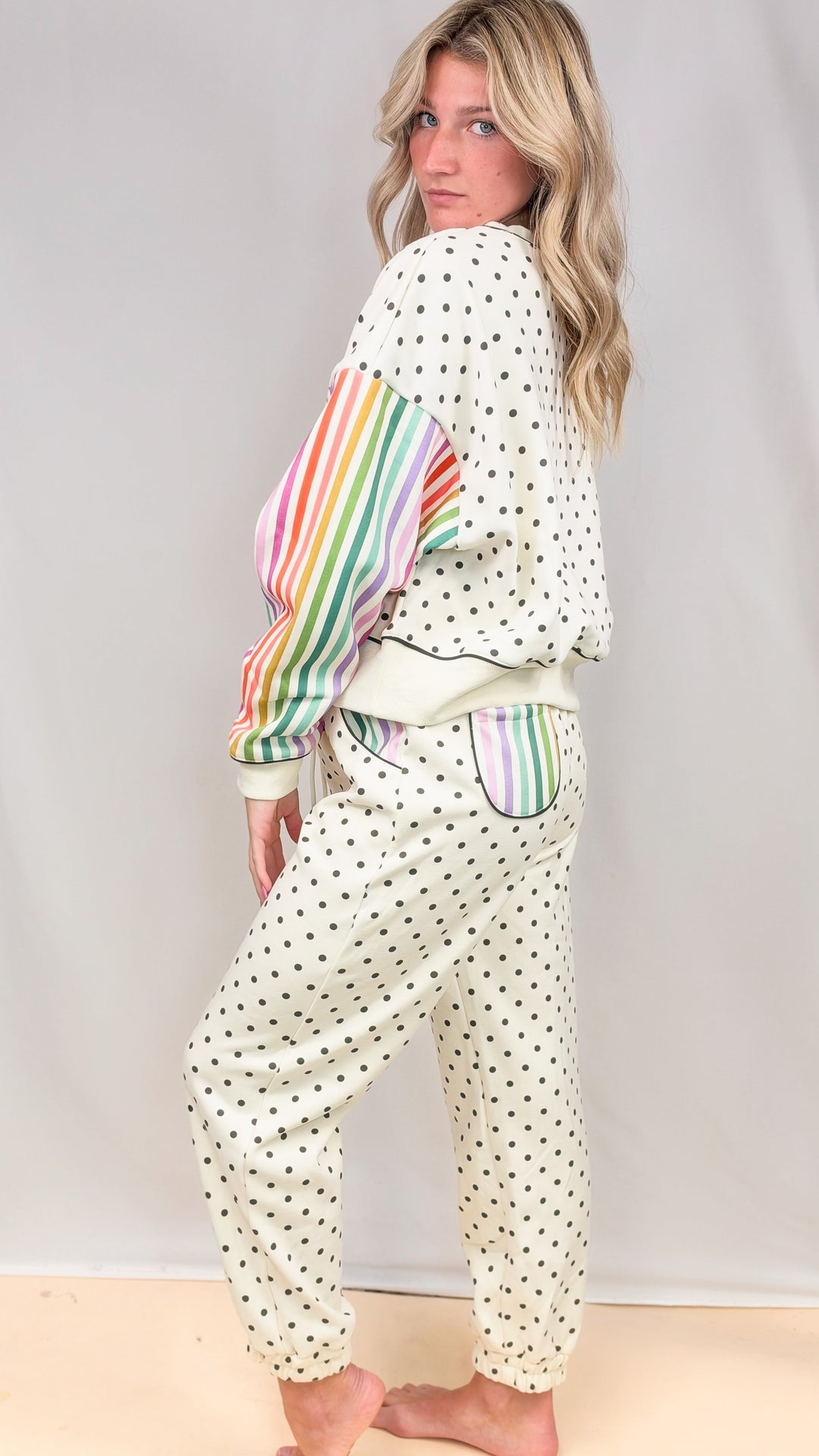 Woman wearing cream matching sweatsuit with rainbow color stripe sleeves and black polka dots on plain background
