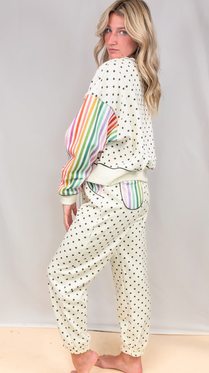 Woman wearing cream matching sweatsuit with rainbow color stripe sleeves and black polka dots on plain background