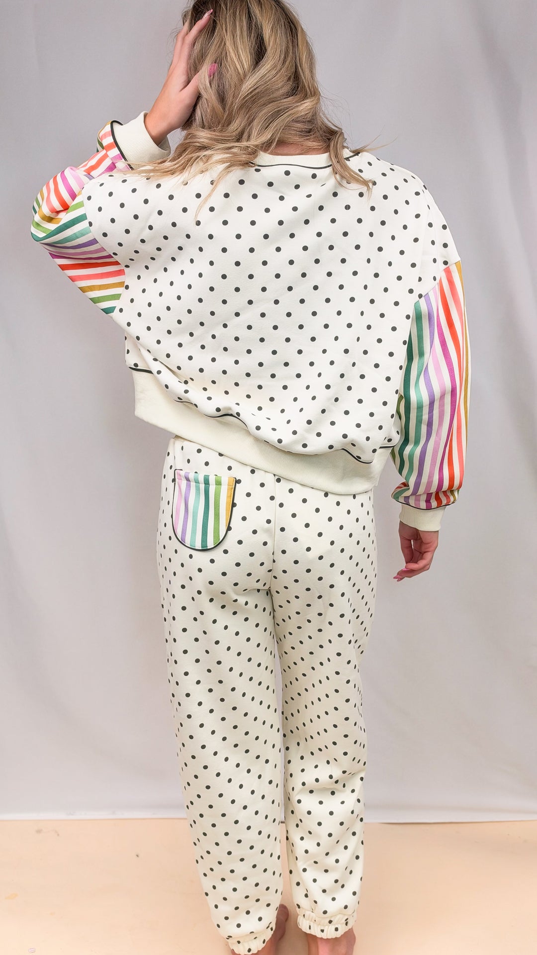 Woman shown from back wearing cream matching sweatsuit with rainbow color stripe sleeves and black polka dots on plain background