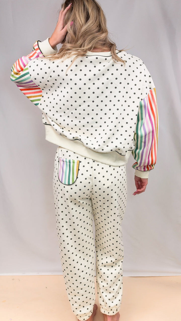 Woman shown from back wearing cream matching sweatsuit with rainbow color stripe sleeves and black polka dots on plain background