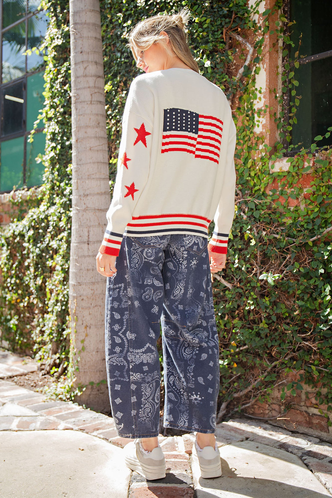 Person wearing a white sweater with an American flag design and patterned pants walking outdoors.