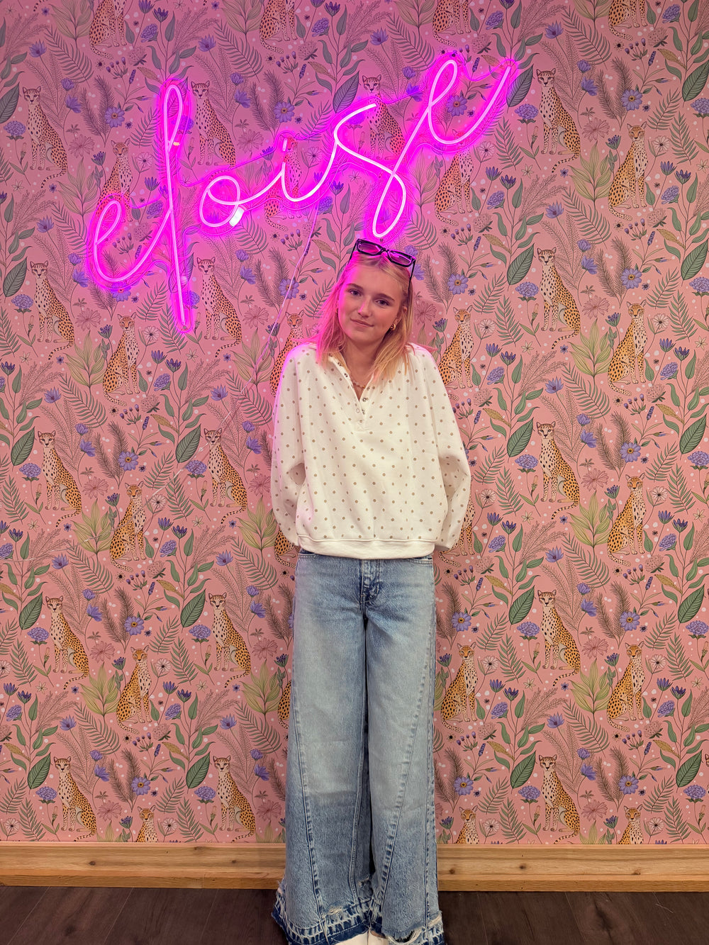 Person standing in front of a pink floral-patterned wall with a neon sign.