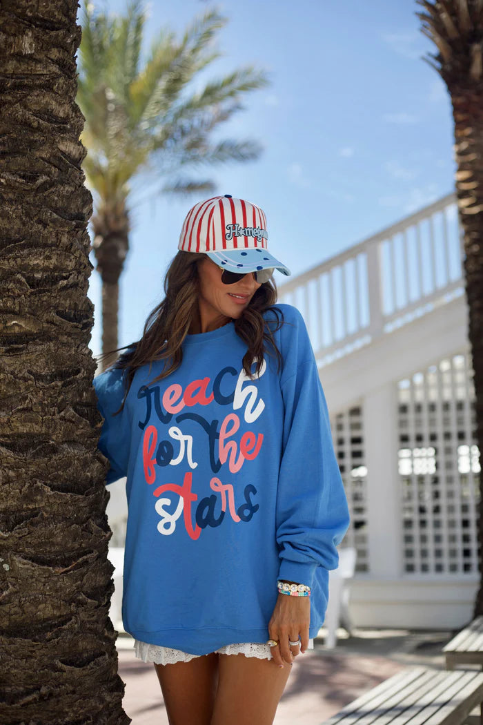 Woman wearing a blue sweatshirt with text and a colorful cap, standing by palm trees.
